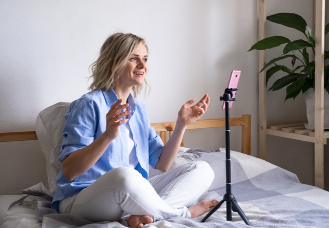 mulher sentada na cama gravando com o celular com a ajuda de um tripé