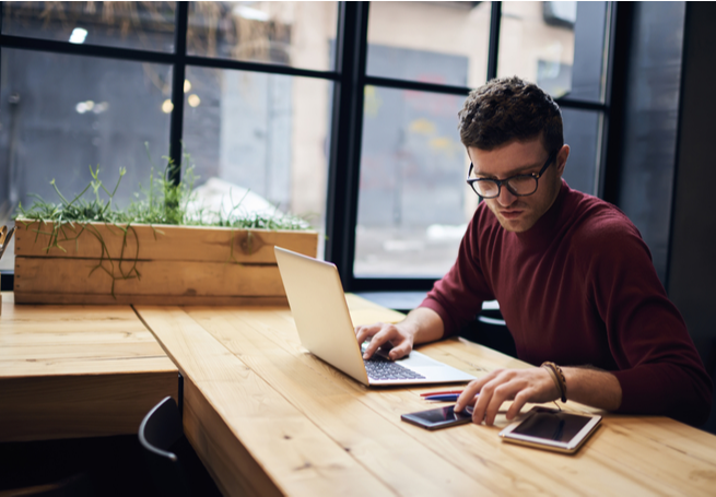 Homem manipulando celular ao lado de um notebook sobre a mesa